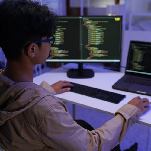 Male Software Engineer Writing Code on Desktop Computer with Multiple Screens Setup. Data center programmer managing large datasets used in AI training procedures to streamline tasks.